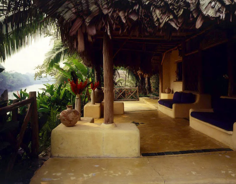 Área de terraza con vista al río Tatín, del mismo ranchón. De nuevo, los sillones están incorporados en la arquitectura, cubiertos por tapicería que utiliza textiles guatemaltecos. El piso es de cemento con pintura amarilla y azulejos, la baranda es en madera de tronco pelado, en estructura cruzada.