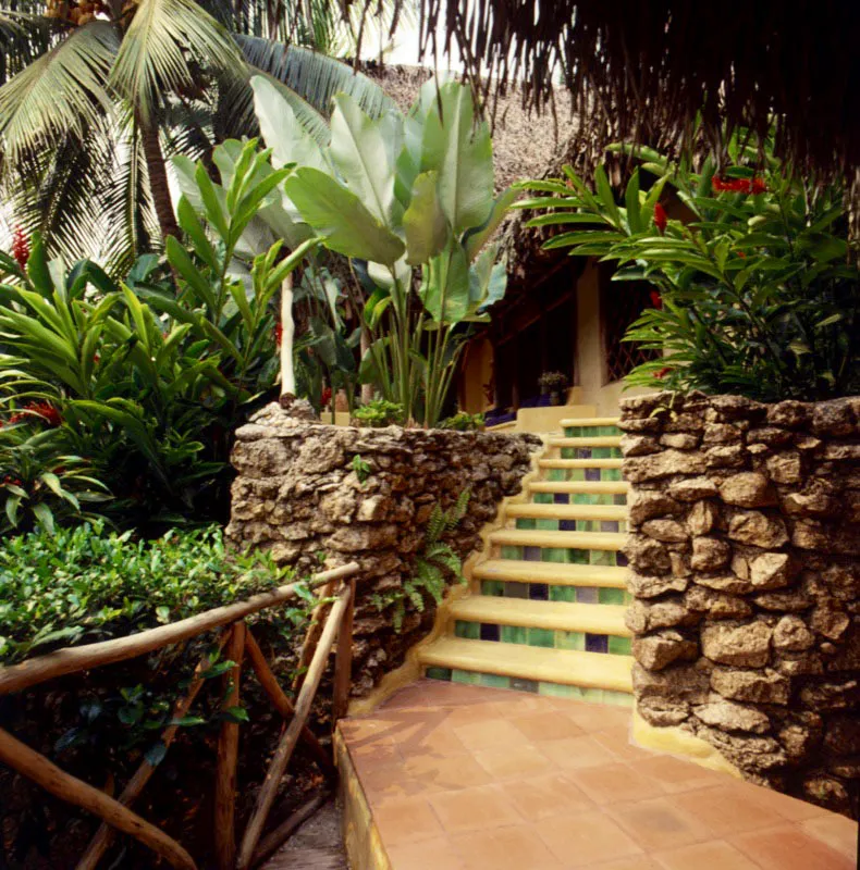 Detalle de una escalera con arriates de piedra a sus lados, en medio de un cuidado jardín con la exuberante vegetación de la región. Se combina la baldosa de barro con escalones de cemento y azulejos.