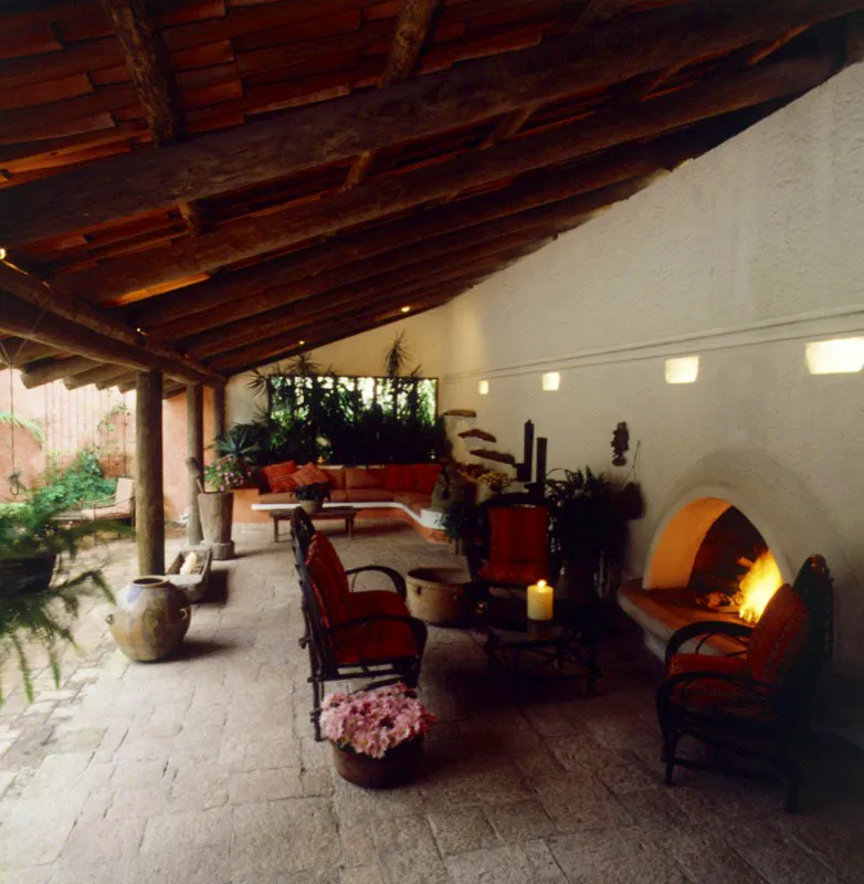 Detalles de una casa contemporánea en ciudad de Guatemala, en la que se usan elementos e ideas de la arquitectura tradicional del país. Una galería exterior con techo de teja y baldosa de piedra; el muro con revoque blanco, sin decoración, salvo la moldura, debajo de la cual hay nichos para luz indirecta, y la chimenea de boca semicircular. Al fondo, una pequeña sala de mampostería, empotrada a la arquitectura. Sobre el piso entre las columnas, un mortero tallado de madera con su mazo, una batea y una olla de barro, todo de origen indígena.