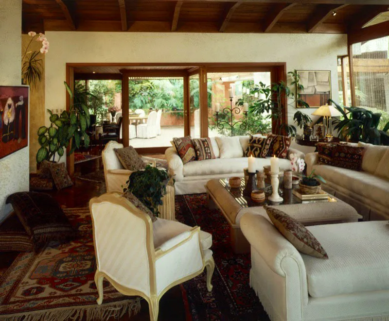 Sala principal de la misma residencia de la ciudad de Guatemala, que se abre al patio jardinizado exterior con amplios ventanales. Sobre varias alfombras orientales se combinan sencillos muebles contemporáneos con otros de tipo tradicional, todos de sobria tapicería blanca, con cojines sobrepuestos fabricados con kilims, que dan el toque de color. En la mesa central de diseño contemporáneo, varias vasijas mayas originales.