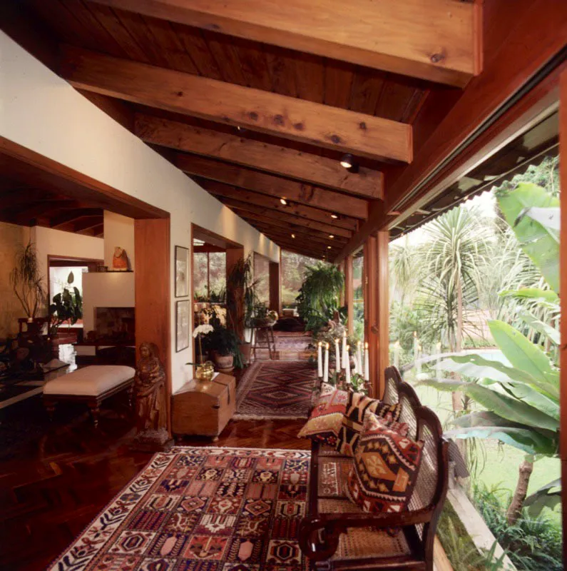 En la misma residencia en ciudad de Guatemala, un pasillo o corredor para circulación entre las salas y los ventanales, en fino parqué de madera, recubierto por alfombras orientales. En primer plano, una banca de madera con petatillo cubierta con cojines de kilim, frente a un santo de madera y un cofre rústico.