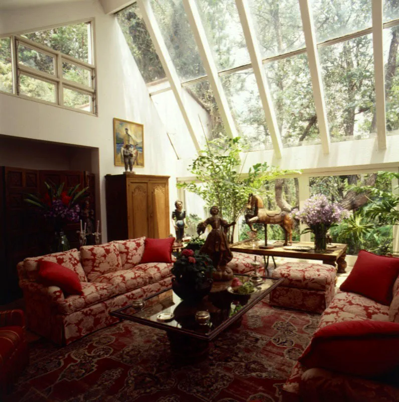 Estar de la misma casa. Se aprecia mejor la amplitud y forma de los ventanales y la vista a los árboles del exterior, así como la excelente alfombra persa y la mesa central sobre la que se exhibe una talla colonial en madera policromada. Detrás, sobre el armario, una figura en terracota, de María Dolores Castellanos, y una pintura del artista guatemalteco Erwin Guillermo. Se aprecia también, sobre una mesa rústica, un antiguo caballo de carrusel de madera encerada.