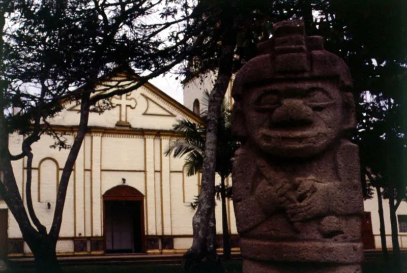 Cultura San Agustín. Figura antropomorfa. Piedra
La colección estatuaria de San Agustín (siglo v d. C.) estuvo destinada al ocultamiento, debido a su naturaleza funeraria. Allí cada pieza representa un personaje específico que se desempeña en el trance del muerto entre la vida pasada y el viaje a una vida futura. Se encuentran figuras de guerreros, sacerdotes y dignatarios, animales simbólicos mezcla de varias zoologías y deidades que combinan en un cuerpo partes felinas y humanas. Esta imagen, proveniente de Ullumbe y ubicada en la Plaza de Bolívar de San Agustín representa a un sacerdote. Los objetos que lleva en la mano –una concha de caracol y un palillo– eran empleados para mezclar la sustancia alcalina y la coca destinadas a las ceremonias rituales, a lo que se suma el tocado de la cabeza, que designa su condición especial.
Lejos de pretender ser una imagen realista, la figura monolítica no guarda las proporciones del ser humano pero está dotada de un vigor grave y vital.