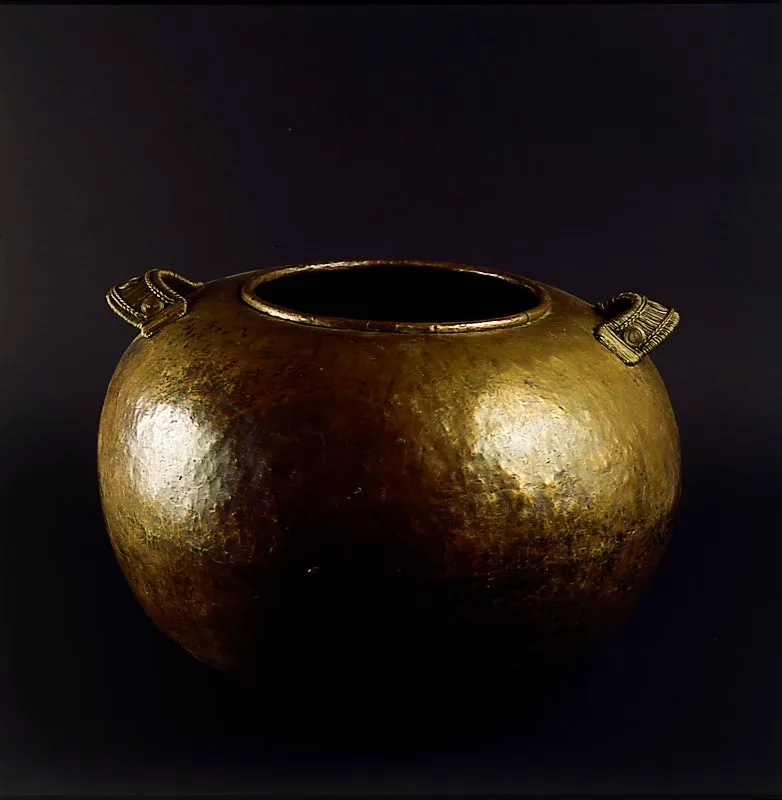Recipiente globular de gran tamaño, en aleación de bronce, ensamblado y martillado, con asas tejidas, ornamentadas y reforzadas con hilo del mismo material. Su forma está inspirada en los recipientes cerámicos prehispánicos. Bogotá. José Fernando Machado
