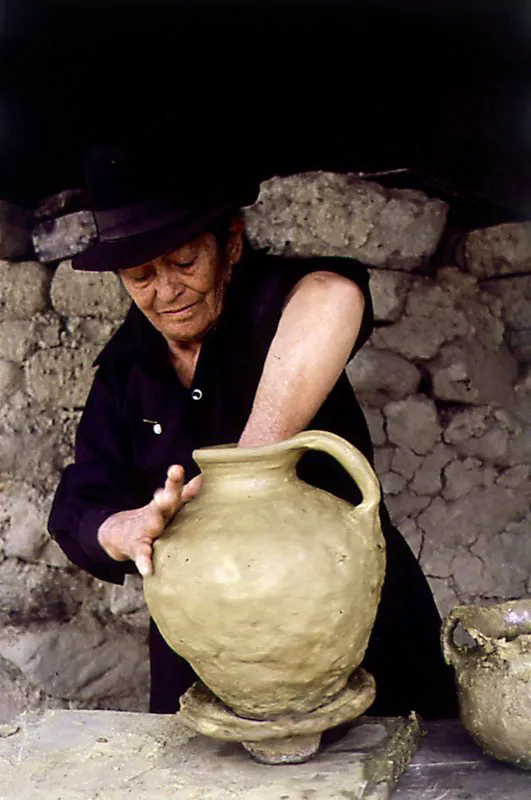 Eva Villanueva elaborando, a mano, un recipiente en barro. El procedimiento consiste en realizar el modelado sobre un plato de bailar la loza, a partir de una bola de barro que se ahueca y que será la pieza que forme el fondo de la vasija. De este primer material se van a levantar las paredes al yuxtaponer nuevo material, en forma de rollos, que al estirar hacia arriba se va adelgazando. Los otros procedimientos tradicionalmente utilizados son el moldeado y el torneado. Ráquira, Boyacá. Diego Miguel Garcés