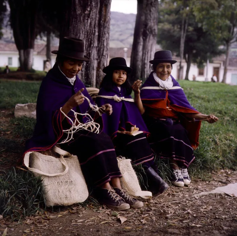 Indias Guambiano en sus trajes tpicos. Una de ellas inicia el tejido de una jigra o mochila en fique, mientras otra enmadeja lana despus de hilarla. Las mujeres visten una serie de anacos o faldas de lana de color negruzco, adornadas con franjas transversales de otro color, que ellas mismas tejen en telar vertical. Las envuelven en su cuerpo asegurndolas a la cintura con una vistosa faja o chumbe tejido en hilo de varios colores y adornado con figuras. Sobre los hombros se colocan no menos de cuatro rebozos azules, de tradicin espaola, cogidos a la altura del pecho con ganchos metlicos. Silvia, Cauca. Alfredo Pinzn