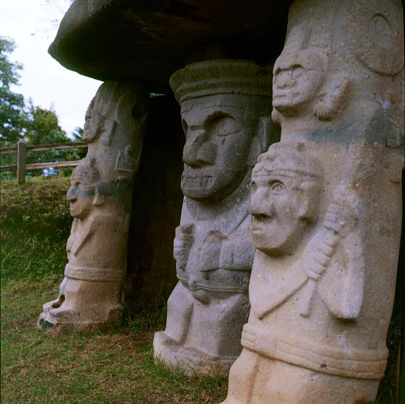 Esta fotografía de las estatuas del Montículo Oriental de la Mesita A del Parque Arqueológico de San Agustín permite apreciar uno de los aspectos formales más notables de la estatuaria. Como puede verse en la escultura central, antes de componer la imagen que se proponía representar, el escultor buscó dar a la piedra forma fálica haciendo elíptico su eje vertical y dando simultáneamente forma cóncava al plano frontal de la estatua al hacer coincidir el extremo del tocado con la punta de los pies. Creaba así un plano de referencia y un eje central con los cuales debía relacionar cada punto, cada línea y cada forma de la futura escultura. El efecto óptico es aún más notable si se tiene en cuenta que la figura está sentada. Algo similar, aunque menos acentuado, se observa en los guerreros que flanquean la estatua.