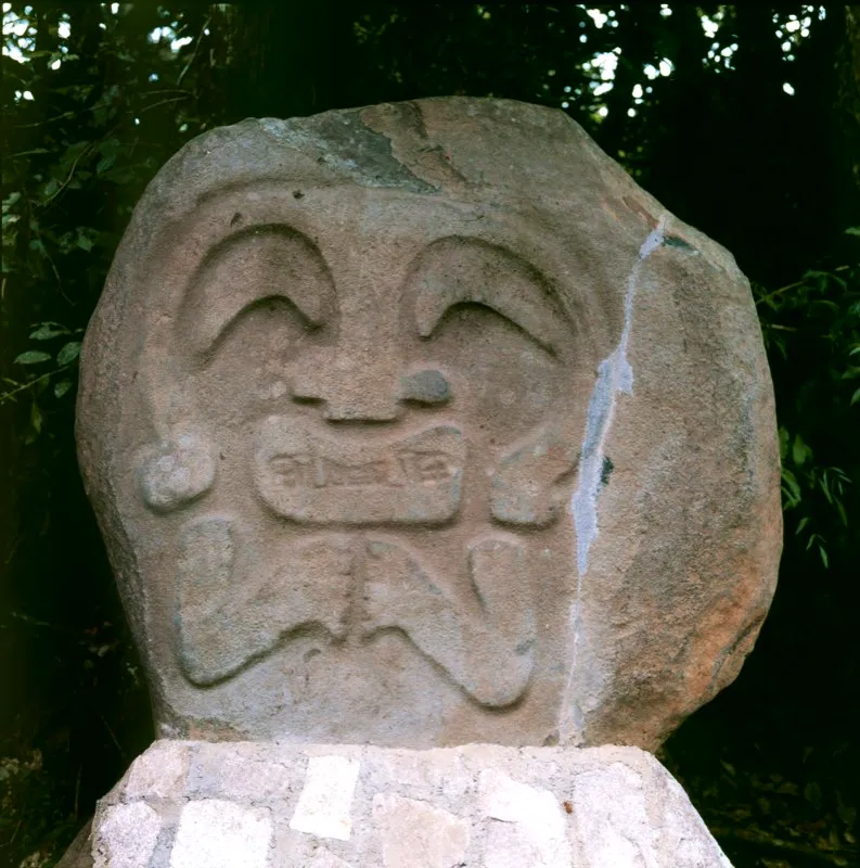 Figura en relieve biplano hallada en Uyumbe, notable porque el escultor solamente resaltó ciertos rasgos en el plano superficial de la piedra: una especie de tocado que enmarca la cara y que se proyecta en el centro para formar la nariz, un par de peculiares ojos en forma de bumerán, las fauces animales y los brazos. El resto lo crea el contemplador en su cerebro, sin que exista en la escultura.