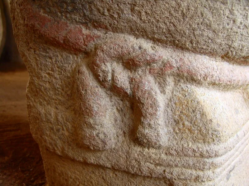 Detalles de la estatua del Montículo V de la Meseta A del Alto de los Ídolos, donde se observan rastros de la pintura que originalmente la cubrió en su totalidad. El tronco y las extremidades estaban pintados de amarillo y de rojo el rostro, el taparrabo con su detallado lazo, y las ligaduras de las piernas. De color castaño estaban pintados su tocado y las cintas que caen por la espalda del personaje.