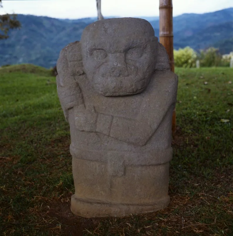 Figura del Alto de Lavapatas, descrita por Preuss como “un guerrero que agarra con ambas manos una maza, rota en el lado superior, y apoyada en el hombro derecho”. Lleva un cinturón del cual “se desprende una faja que cubre las partes pudendas”, además de una cinta en la cabeza, orejeras de tres anillos y brazaletes en ambas muñecas, indumentaria humana que contrasta con los claros rasgos animales de su rostro.