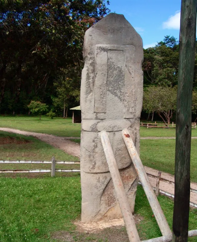 Una de las imágenes más extraordinarias de la estatuaria del Macizo Colombiano es la llamada “El Obispo”, del Montículo Noroeste de la Mesita B del Parque Arqueológico de San Agustín, vista aquí por su cara posterior. Es una estatua de doble faz, que por este lado presenta la espalda de una figura probablemente femenina, provista de una especie de faldellín y una cinta que cuelga de su cabeza. Por su cara anterior muestra algo muy distinto. Los soportes de hierro que la sostienen en su sitio son la culminación de una historia de incomprensión de esta estatua. Describiéndola como “un magnífico fracaso”, el arqueólogo José Pérez de Barradas no podía entender cómo el escultor omitió la base para hincarla en el suelo. Sería imposible —pensó— que la estatua “estuviera la mitad en el aire y la mitad enterrada”.