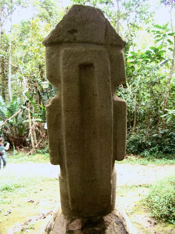 Estatua del Montículo Noroeste de la Mesita B del Parque Arqueológico de San Agustín vista por su plano posterior. En esta figura, que se encuentra sentada en el banco alto propio de la estatuaria, las cintas cuelgan desde el tocado piramidal que parece identificar la imagen del sol y se prolongan hasta los talones. Es un aditamento que comparten las imágenes de chamanes con figuras más sagradas.