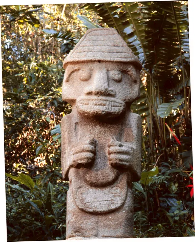 Estatua del Montículo Noroeste de la Mesita B del Parque Arqueológico de San Agustín vista de frente. En esta figura, que se encuentra sentada en el banco alto propio de la estatuaria, las cintas cuelgan desde el tocado piramidal que parece identificar la imagen del sol y se prolongan hasta los talones. Es un aditamento que comparten las imágenes de chamanes con figuras más sagradas.