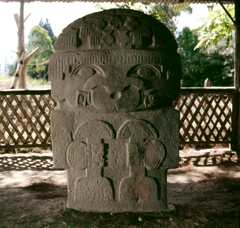 Estatua hallada en Quebradillas, una de las tres conocidas que muy probablemente representan escultores, y de las tres la más ornamentada. Una nariguera en forma de medialuna cubre sus grandes fauces animales, dejando ver con claridad los colmillos, y las muñecas están adornadas con brazaletes de varias vueltas. El tocado semiesférico muestra por su parte anterior cinco figuras de aves de presa en síntesis geométrica y en posición invertida, y franjas estriadas que se repiten en la parte posterior y forman el tocado o peinado en forma de pirámide escalonada frecuente en la estatuaria. El ave de presa, con su característico pico encorvado, aparece de nuevo en el dibujo de los ojos. En las manos lleva dos instrumentos idénticos que el primero en describir la estatua, José Pérez de Barradas, vio como “dos cetros (?) o instrumentos”.